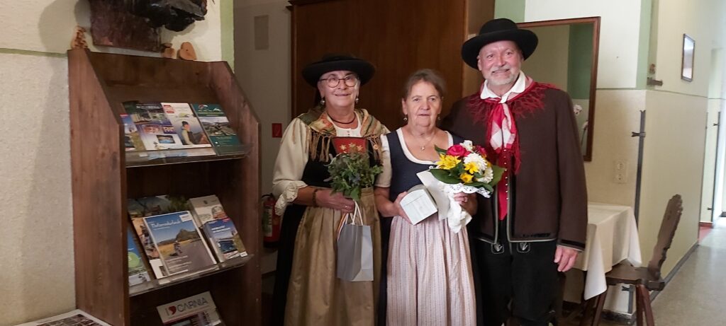 Trachtengruppe mit Blumen und Geschenken vor einem Regal mit Prospekten